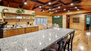 Una cocina amplia con encimeras de granito, armarios de madera y una isla grande. Hay varias sillas alrededor de la isla, y el techo presenta vigas de madera a la vista e iluminación empotrada.
