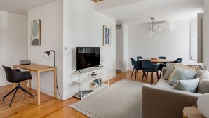 Ett modernt vardagsrum med trägolv, en platt-TV på ett vitt TV-bord, en matplats med bord och stolar samt en grå soffa.
