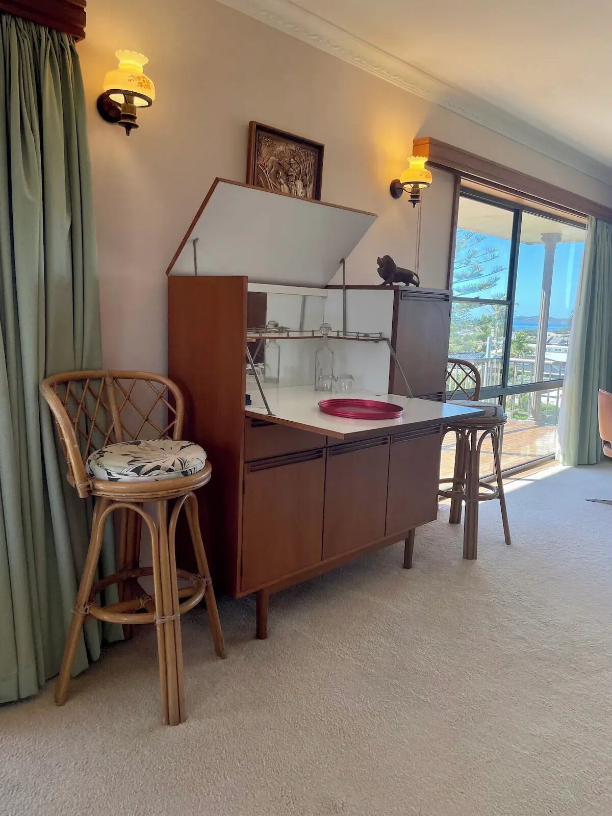 Une pièce avec un bureau en bois, deux lampes fixées au mur, une chaise avec un coussin et une fenêtre donnant sur des palmiers.