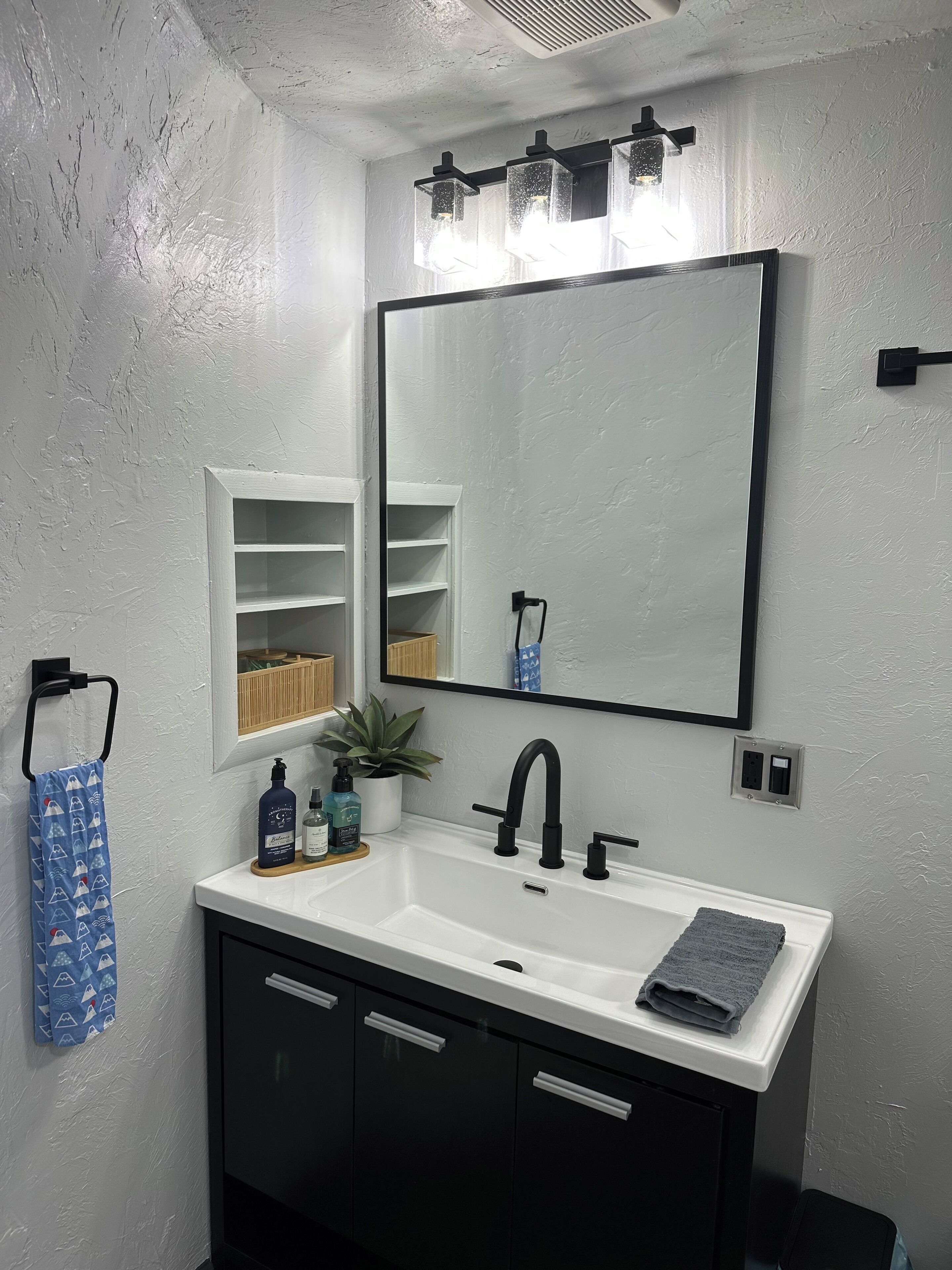 A bathroom with a white sink, black faucet, and a large mirror above it. There is a towel rack with a blue towel and a small shelf with toiletries.