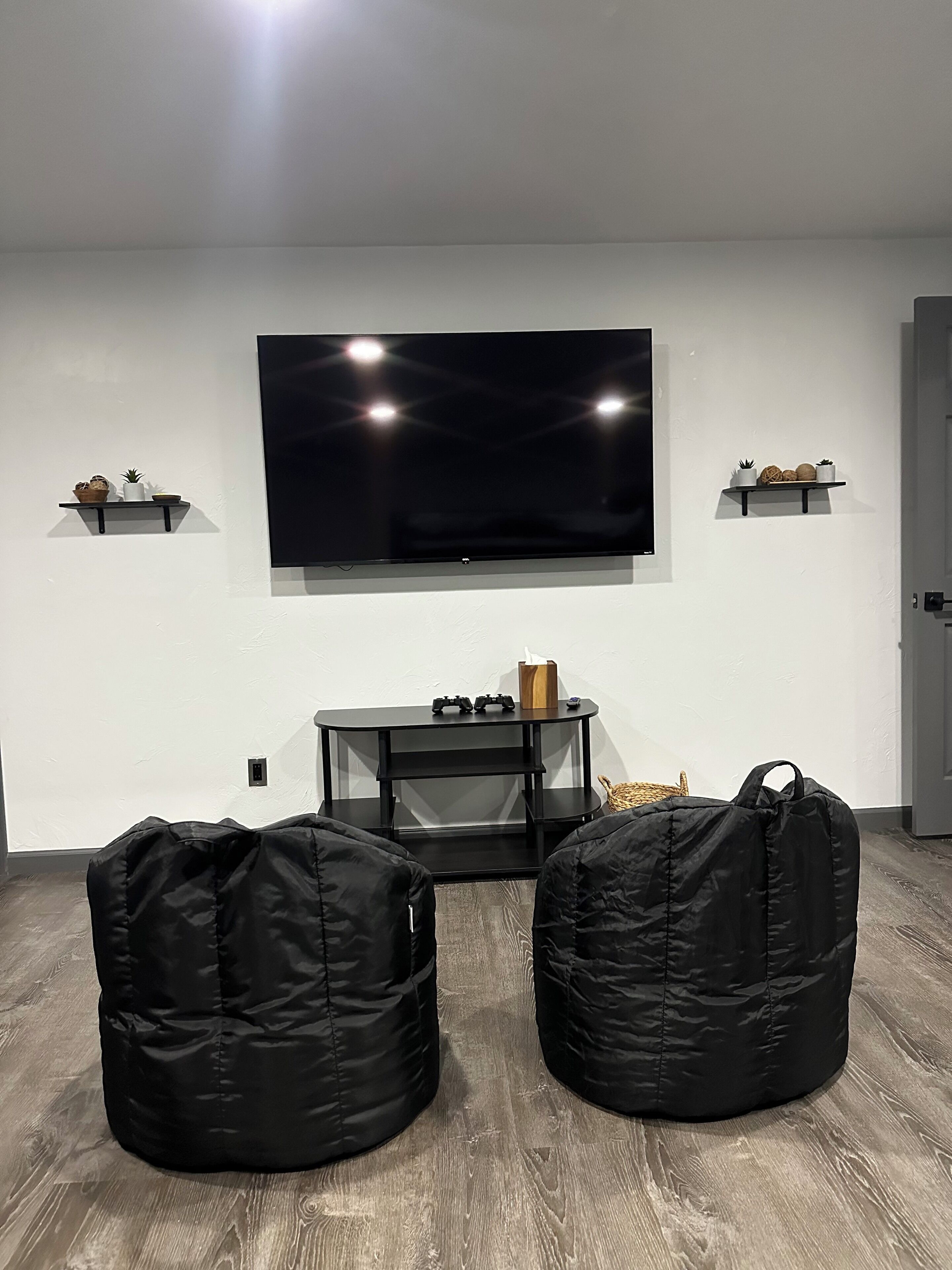 Uma sala de estar com televisão de tela plana na parede, dois pufes, um rack preto e prateleiras com objetos decorativos.