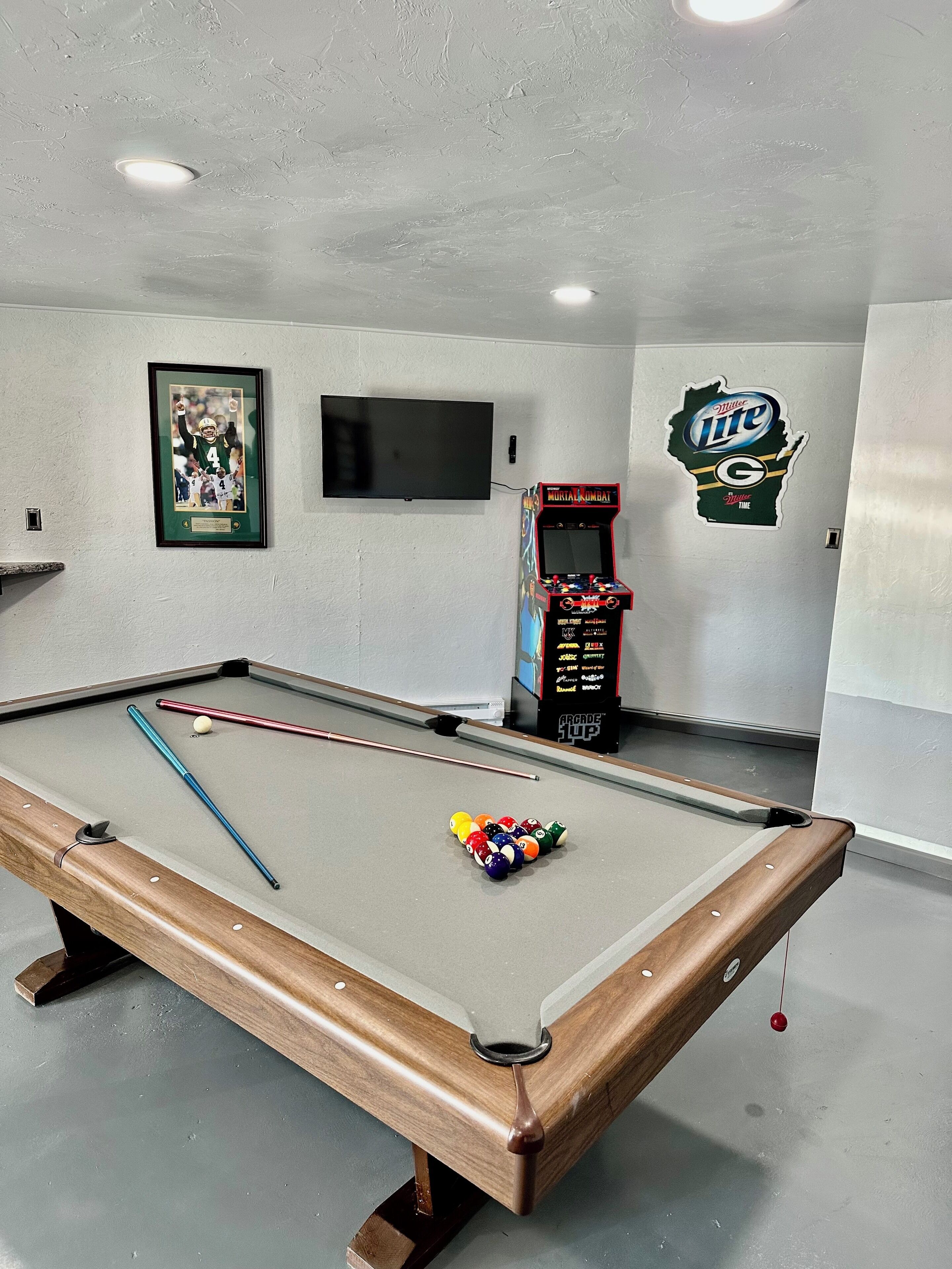 A pool table with billiard balls and cues, a framed sports poster, a television, and a Miller Lite arcade machine in a room with white walls.