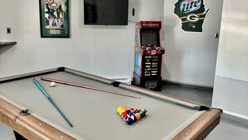 A pool table with billiard balls and cues, a framed sports poster, a television, and a Miller Lite arcade machine in a room with white walls.
