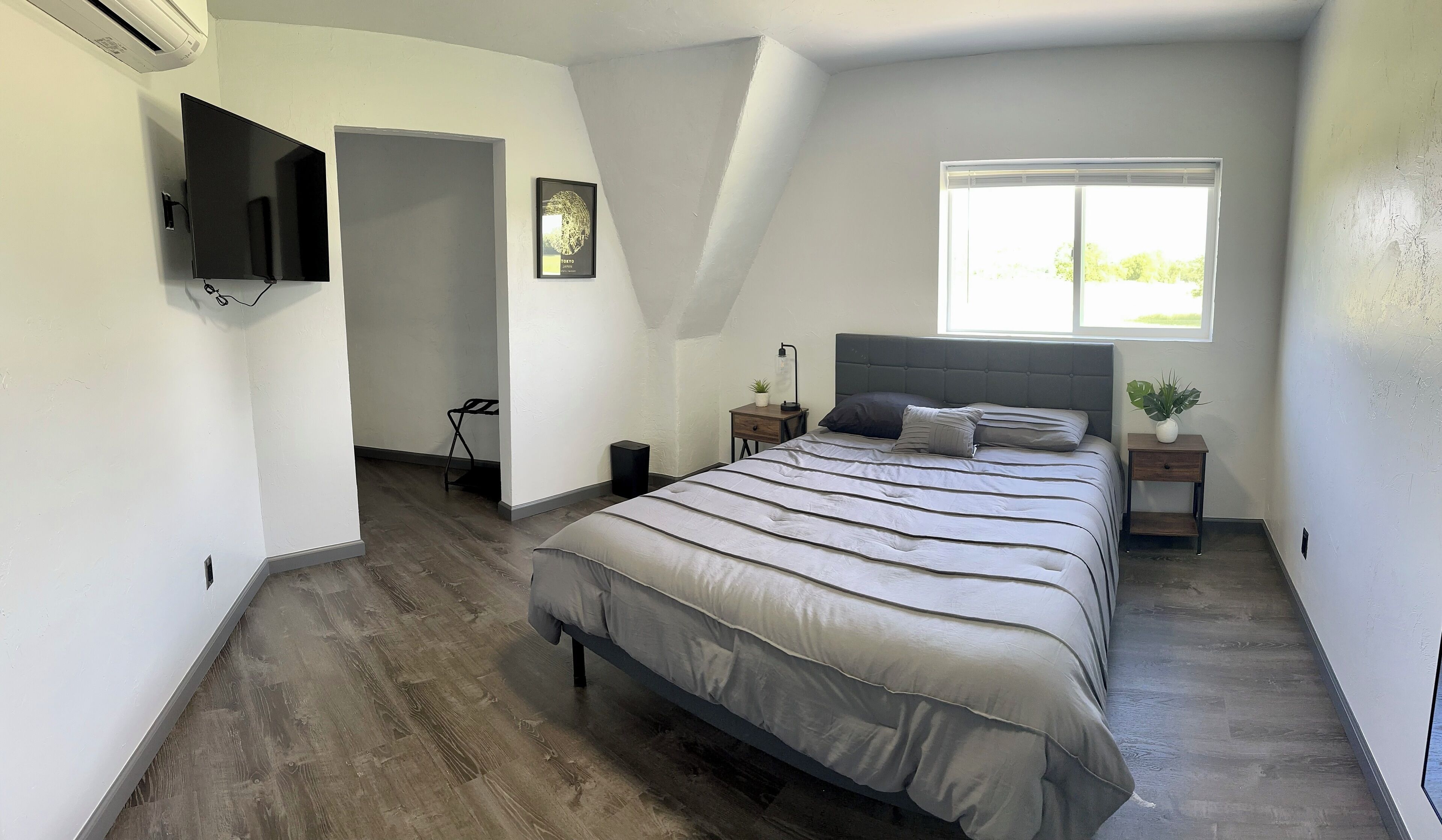 Ein Schlafzimmer mit einem großen Bett, einem an der Wand befestigten Fernseher, einem Fenster und einem kleinen Tisch mit einer Pflanze.