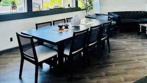 A dining area with a large table, chairs, a chandelier, and a view of trees through large windows.
