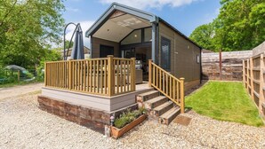 A single-story building with a covered porch, a wooden deck, and a gravel driveway. - Woods Meadow Glamping (West Malling)