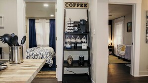Eine Küchenzeile mit einer Kaffeestation, einem Esstisch mit Metallbehälter und einem Bett mit gemusterten Betttextilien.