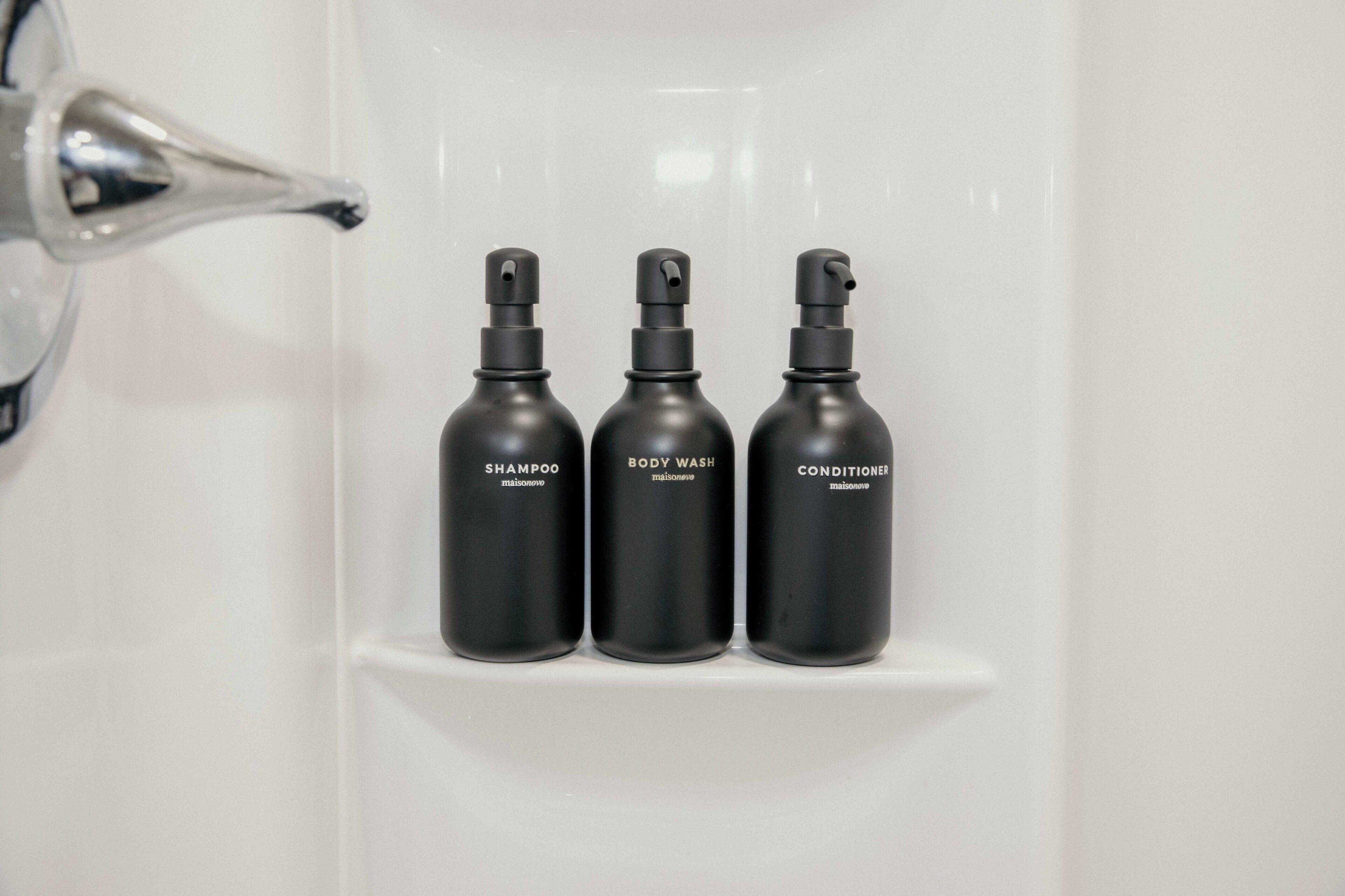 Trois bouteilles noires étiquetées « shampoing », « gel douche » et « après-shampoing » sont posées sur une étagère blanche.