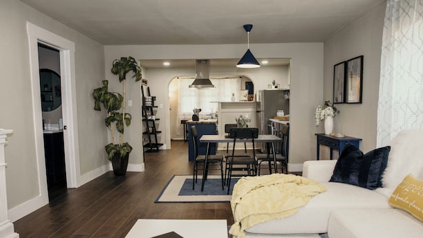 Ein modernes Wohnzimmer mit einem weißen Sofa, einem Couchtisch und einem Essbereich mit Tisch und Stühlen. Im Hintergrund ist eine Küche mit Herd und Schränken zu sehen.