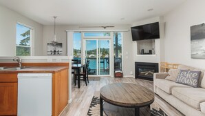 A modern kitchen with a dishwasher, dining area with chairs, a round wooden table, a sofa with a pillow, a fireplace, and a TV mounted on the wall. - Lakefront Townhome with Dock on Devils Lake (Lincoln City)