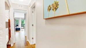 A hallway with wooden flooring, white walls, and a door leading to a room with a bed and a window. - Community Pool | Beach Condo With Resort Amenities (Tybee Island)