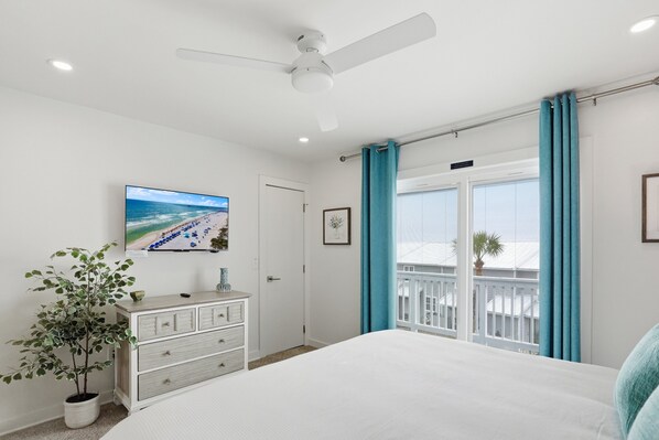 A bedroom with a bed, a dresser, a ceiling fan, a TV, a window with curtains, and a plant.