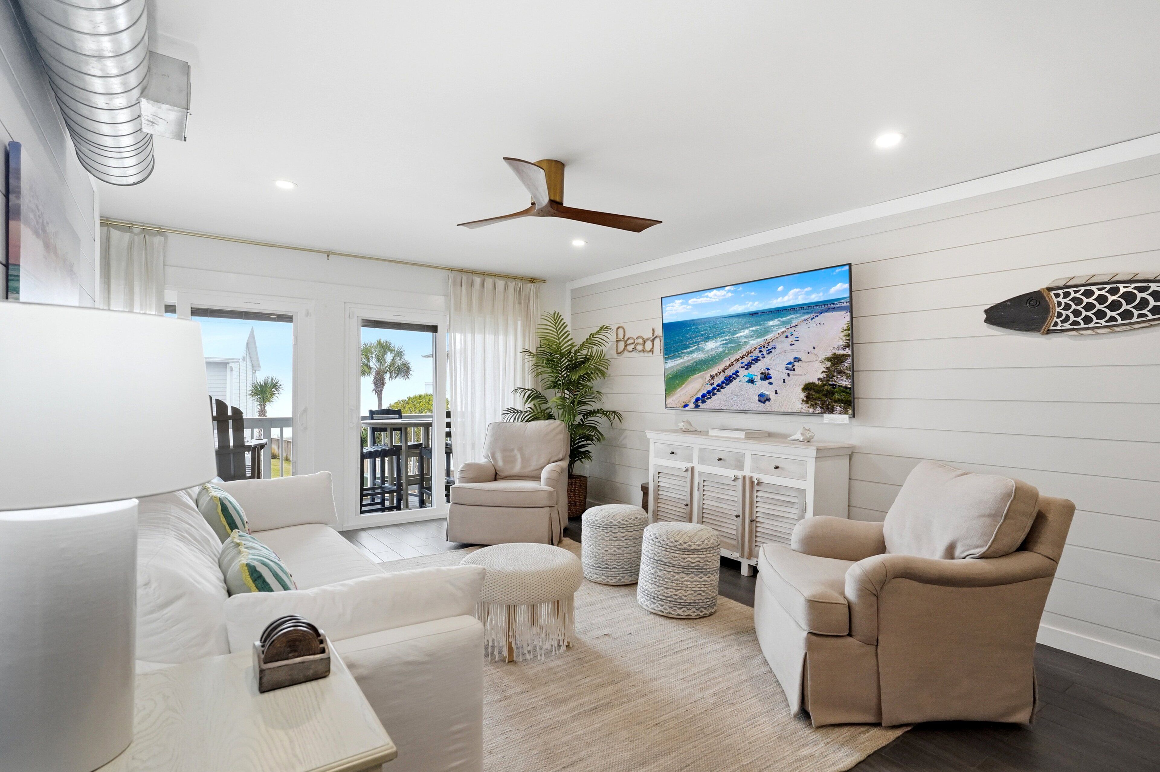 A living room with a sofa, ottomans, a ceiling fan, a TV, and a beach-themed wall art.