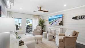A living room with a sofa, ottomans, a ceiling fan, a TV, and a beach-themed wall art. - Shoreline Breeze | Steps to the Beach | Beautiful views from 2nd and 3rd balconies (Inlet Beach)
