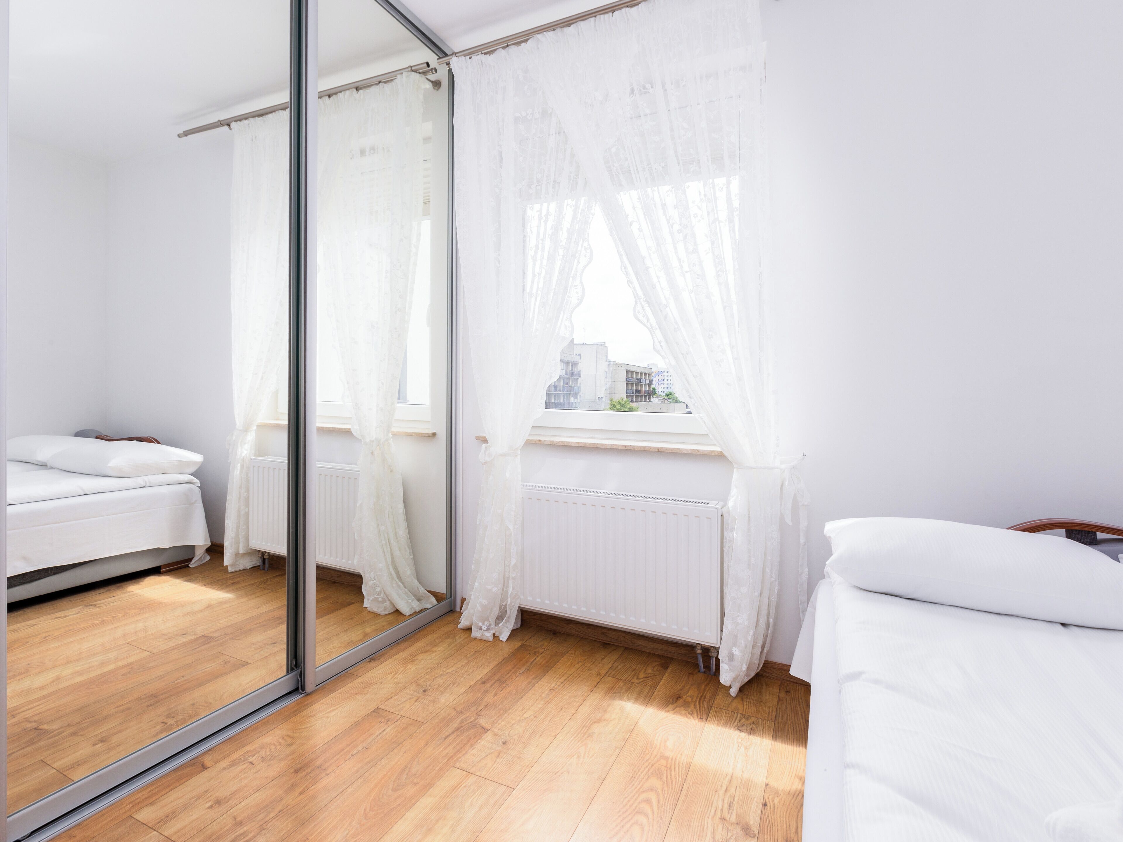 A bedroom with a large mirror, a bed, a radiator, and a window with sheer curtains.