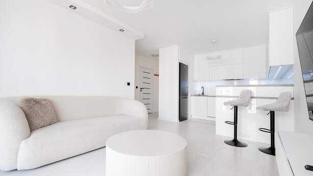 Un salon moderne et minimaliste, avec un canapé blanc, une table basse ronde et un coin cuisine doté de meubles blancs et d’un réfrigérateur noir.