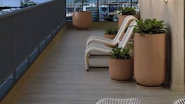 Un balcón con sillas de mimbre y plantas en macetas, con vista a una masa de agua y al paisaje urbano.