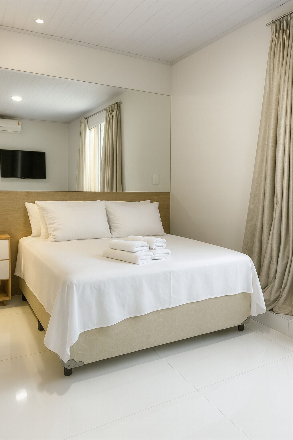 Un lit bien fait, avec du linge de lit et des serviettes blanches, une table de chevet, un téléviseur fixé au mur et un grand miroir sur le mur.