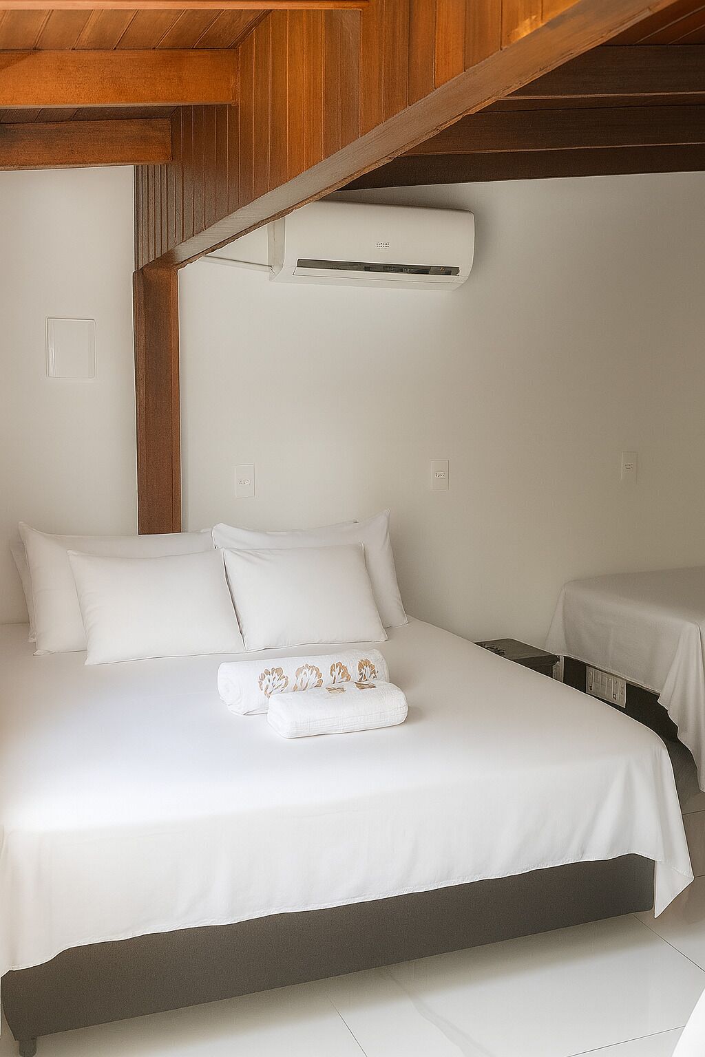 A hotel room with a bed, a bedside table, a small table, and an air conditioning unit mounted on the wall.