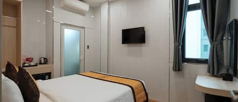 A modern hotel room with a bed, a TV mounted on the wall, a small desk with a coffee maker, and a window with curtains.