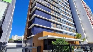 Un bâtiment moderne à plusieurs étages, doté d’une façade combinant verre et bois, et caractérisé par une entrée imposante avec une porte en verre et un petit espace extérieur agrémenté de plantes.