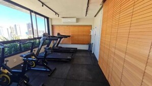 Une salle de sport équipée de tapis de course et de vélos d’appartement, de grandes fenêtres offrant une vue sur la ville et d’un store en bois sur le côté droit.