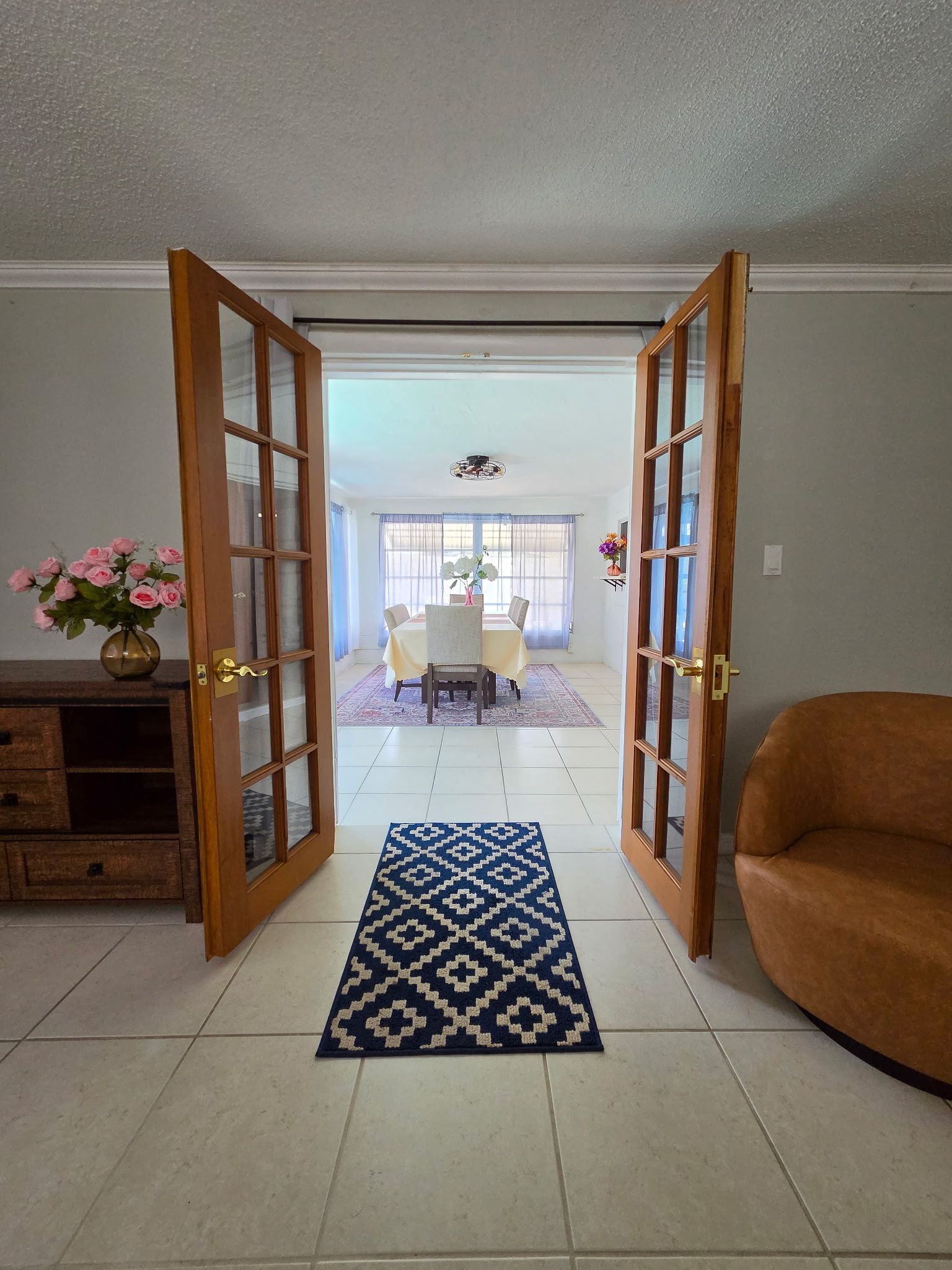 A room with a dining table, chairs, a blue patterned rug, a brown sofa, and a wooden cabinet with a vase of flowers.