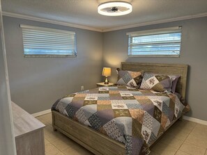 Cozy retreat with a quilted bedspread and a wooden bed frame, featuring a nightstand with a lamp and two windows with blinds.