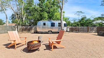A gravel area with two wooden chairs and a metal fire pit. A silver RV is parked in the background.