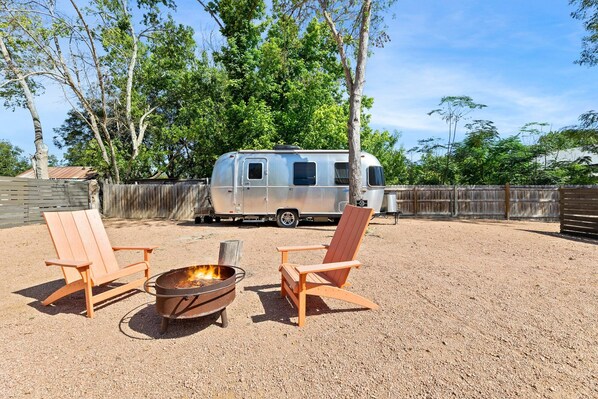 A gravel area with two wooden chairs and a metal fire pit. A silver RV is parked in the background.
