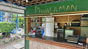 Um pequeno quiosque de comida chamado "PiWALAMAN", com balcão, utensílios pendurados e uma geladeira.