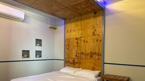 Quarto individual com uma cama de madeira, um pequeno banco de madeira e duas prateleiras fixadas na parede com objetos decorativos.