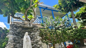Estátua de uma figura vestida em um pedestal de pedra em um jardim, com um edifício ao fundo.