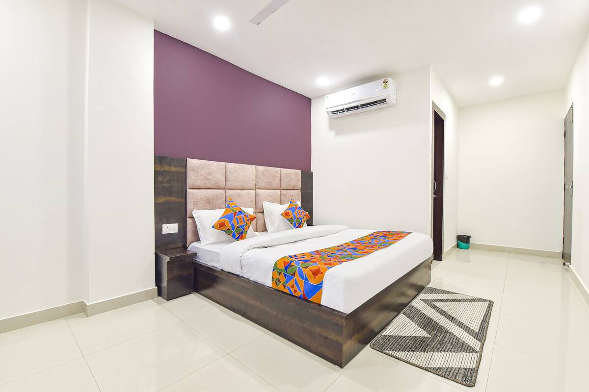A modern hotel room with a large bed, a headboard, a bedside table, an air conditioning unit, and a bathroom door.