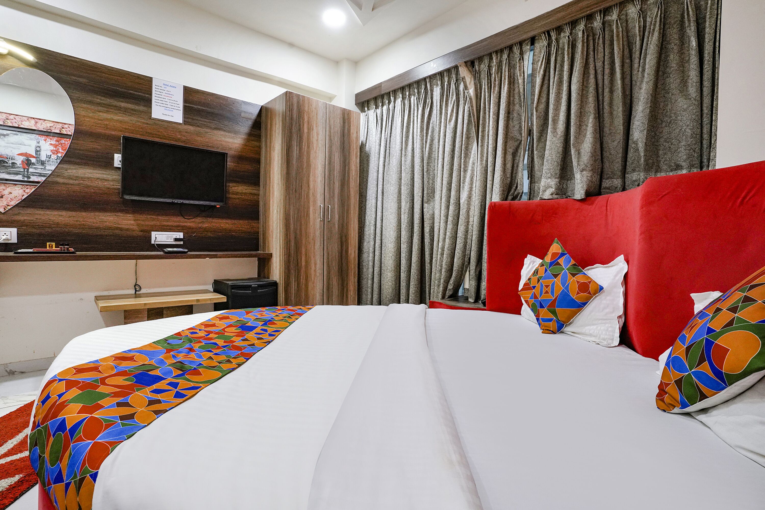 A hotel room with a large bed, a TV mounted on the wall, a wooden panel design, and a red headboard.