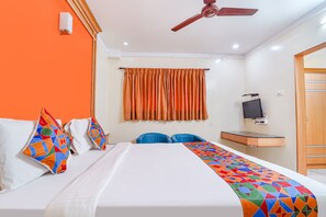 Habitación de hotel con una cama, un escritorio con computadora, un ventilador de techo y una ventana con cortinas anaranjadas.