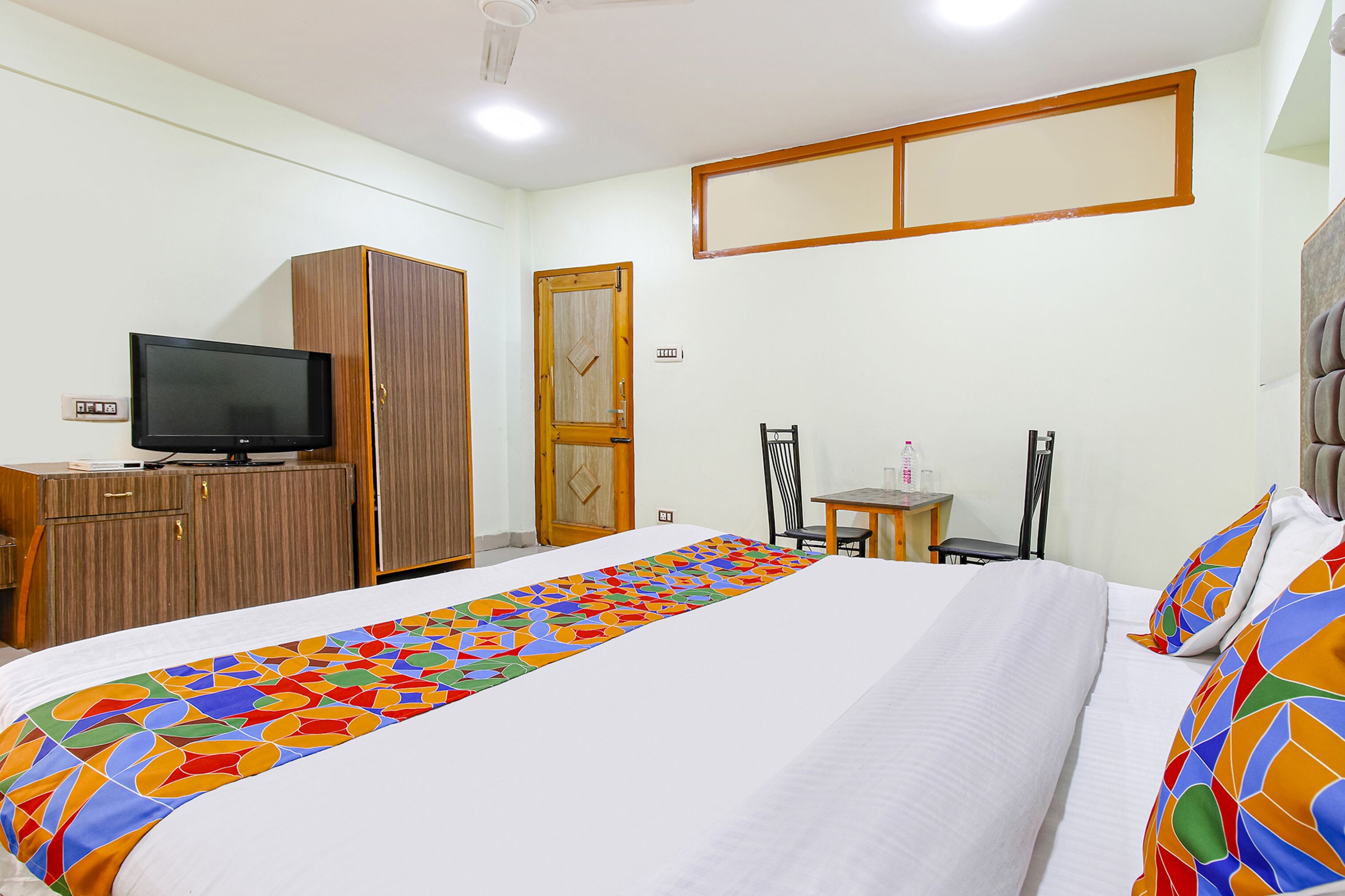 A hotel room with a bed, a television on a wooden cabinet, a small table with a chair, and a wooden door.