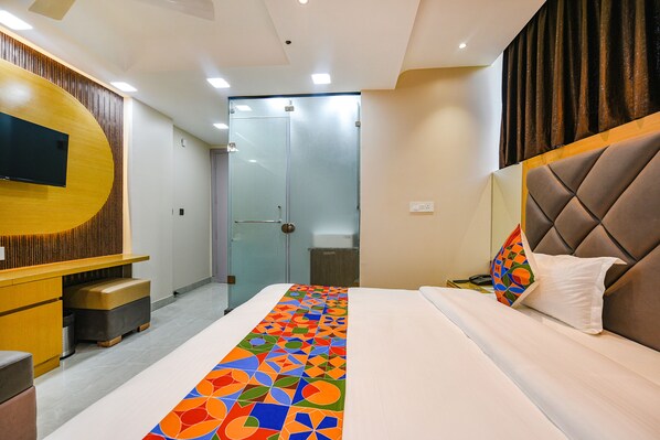 Une chambre d’hôtel avec un lit, un téléviseur fixé sur un panneau en bois, une porte en verre et une salle de bain visible à travers la porte.