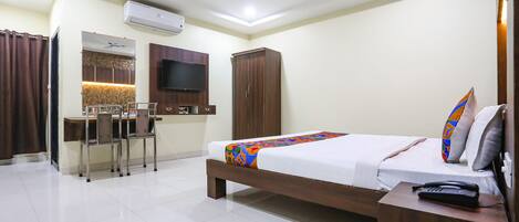 A hotel room with a bed, a desk with chairs, a television, an air conditioner, and a ceiling fan.