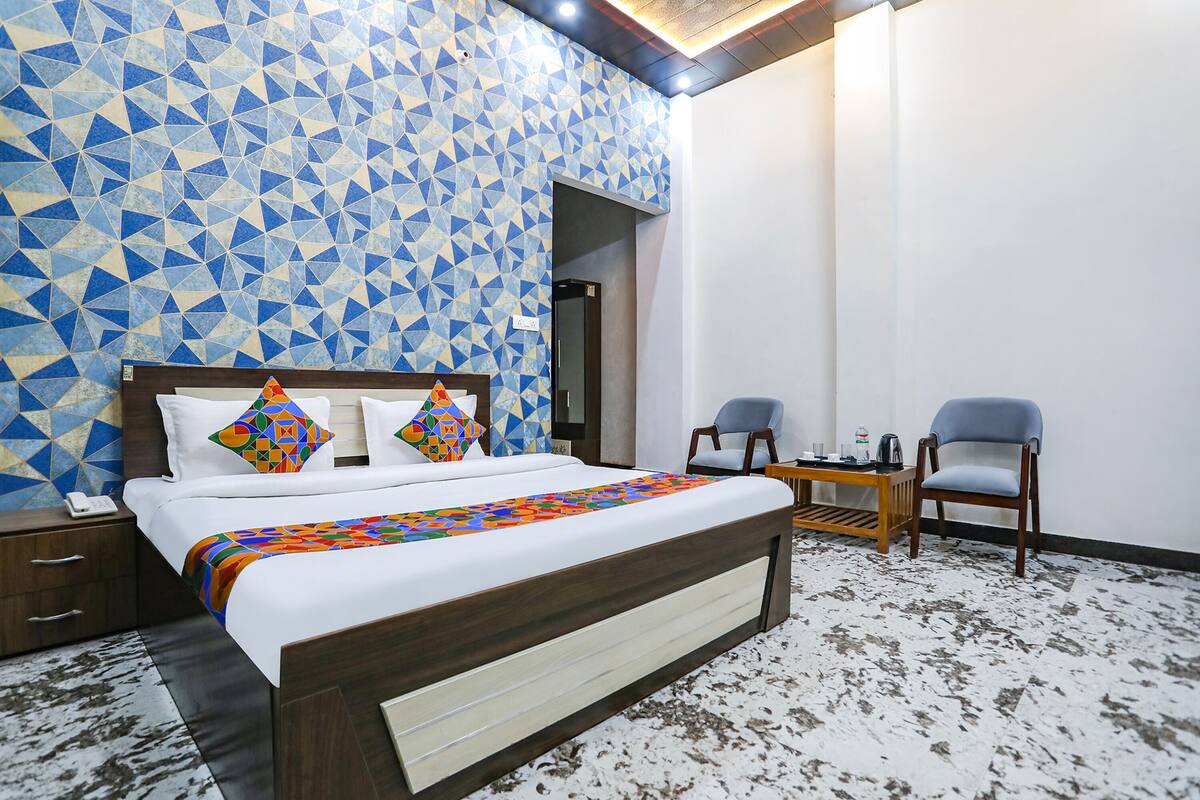 Une chambre d’hôtel avec un mur orné d’un motif géométrique, un lit en bois, deux chaises et une petite table sur laquelle se trouve une bouilloire.