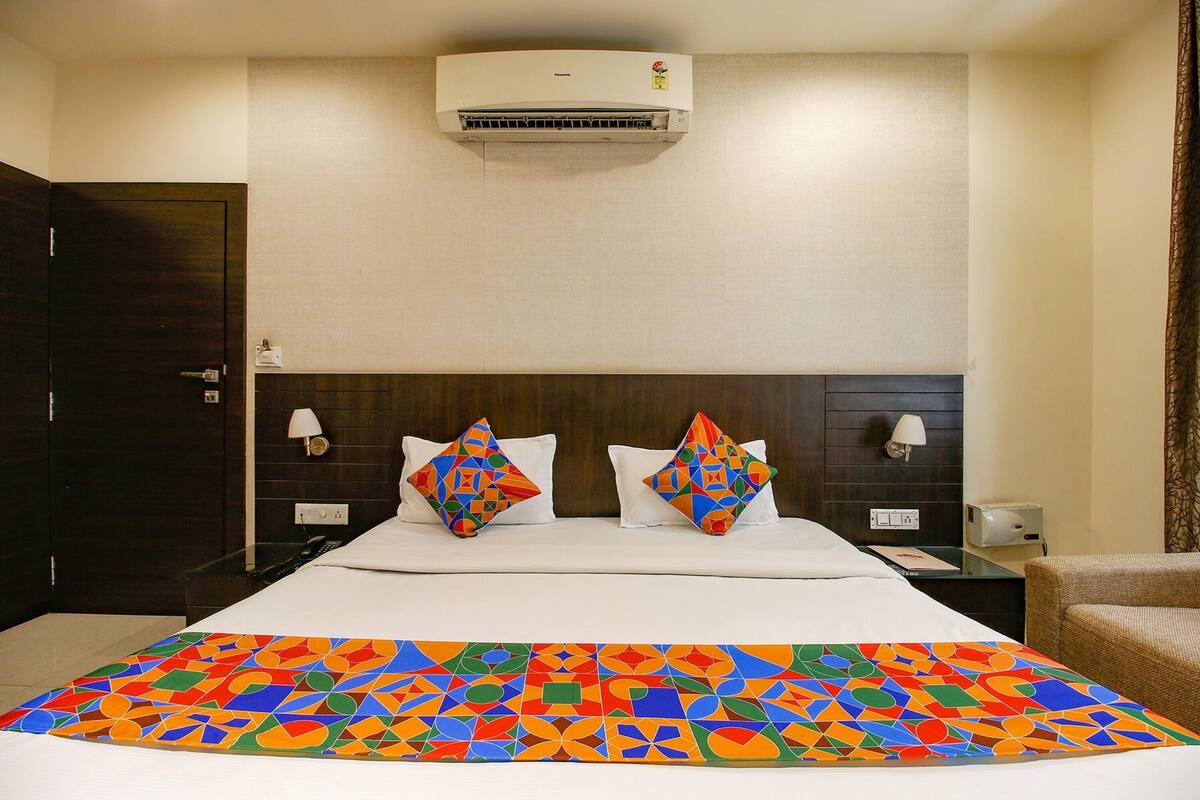 A hotel room with a bed, two pillows, a colorful bedspread, a nightstand, a lamp, an air conditioner, and a chair.