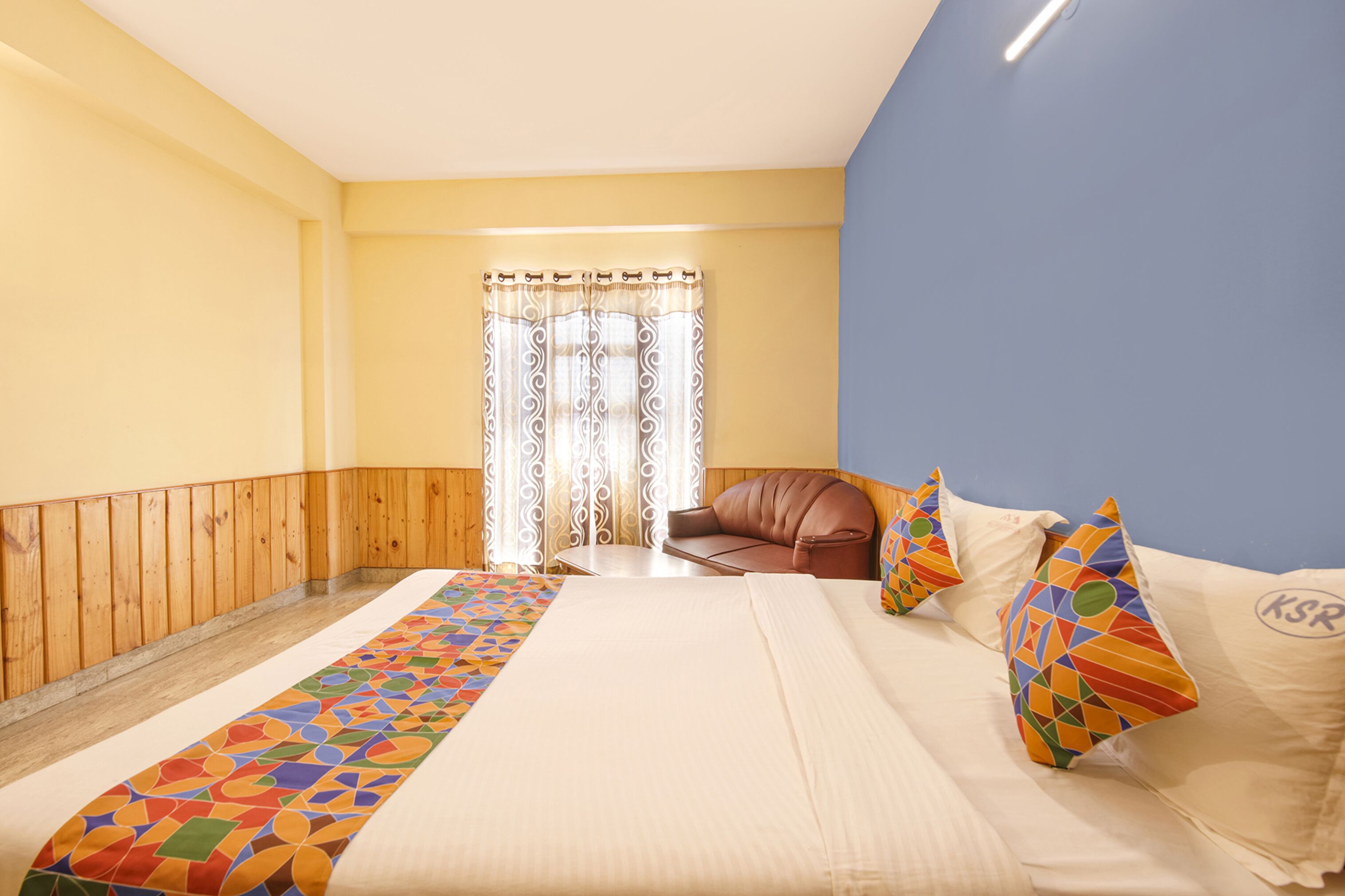 Ein Hotelzimmer mit einem Bett, einem braunen Ledersessel, einem farbenfrohen Teppich und einem Vorhang mit geometrischem Muster.