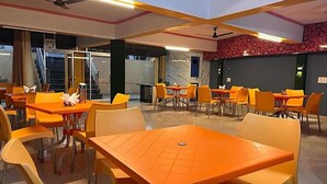 A dining area with orange tables and chairs, a ceiling fan, and a staircase in the background. - Fabhotel New Raghukul Loung (Faizabad)