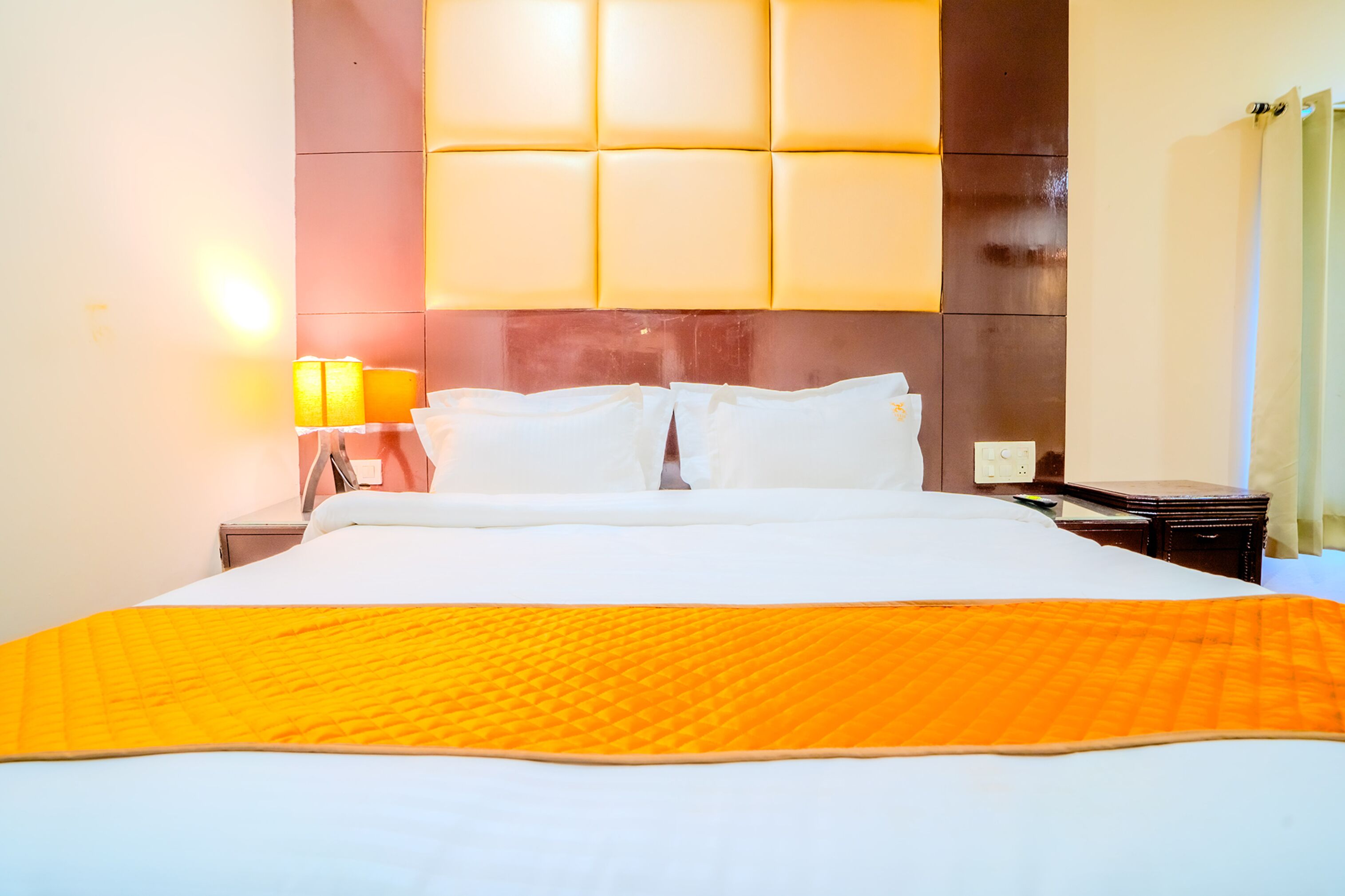 A hotel room with a large bed, a headboard with a light panel, a nightstand with a lamp, and a wooden side table.