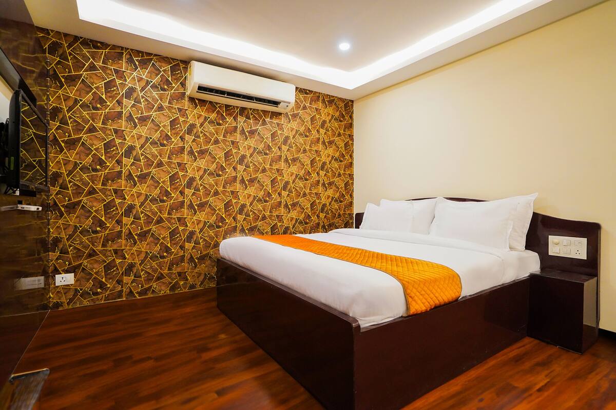 Une chambre d’hôtel moderne avec un plancher en bois, un lit recouvert de draps blancs et agrémenté d’un plaid orange, une table de chevet et un mur orné d’un motif géométrique.