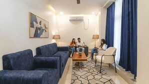 A living room with a sofa, a coffee table, and a chair. There is a ceiling fan, an air conditioning unit, and a painting on the wall. - Fabhotel Raj Palace (Lucknow)
