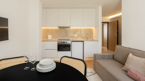 Una cocina moderna con armarios blancos, horno empotrado y encimera con fregadero. Hay una mesa de comedor con platos y vasos, y un sofá en la sala de estar.