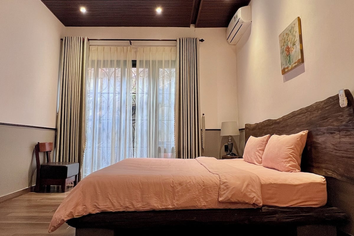 Ein Schlafzimmer mit einem Bett, einem Nachttisch, einer Lampe, einem Fenster mit Vorhängen und einer Klimaanlage.
