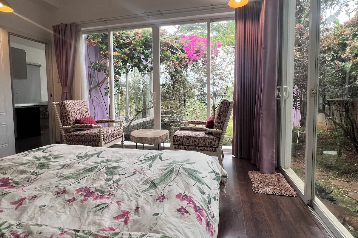Ein Schlafzimmer mit einem großen Bett, zwei Stühlen, einem kleinen Tisch und Blick auf einen Garten durch große Fenster.