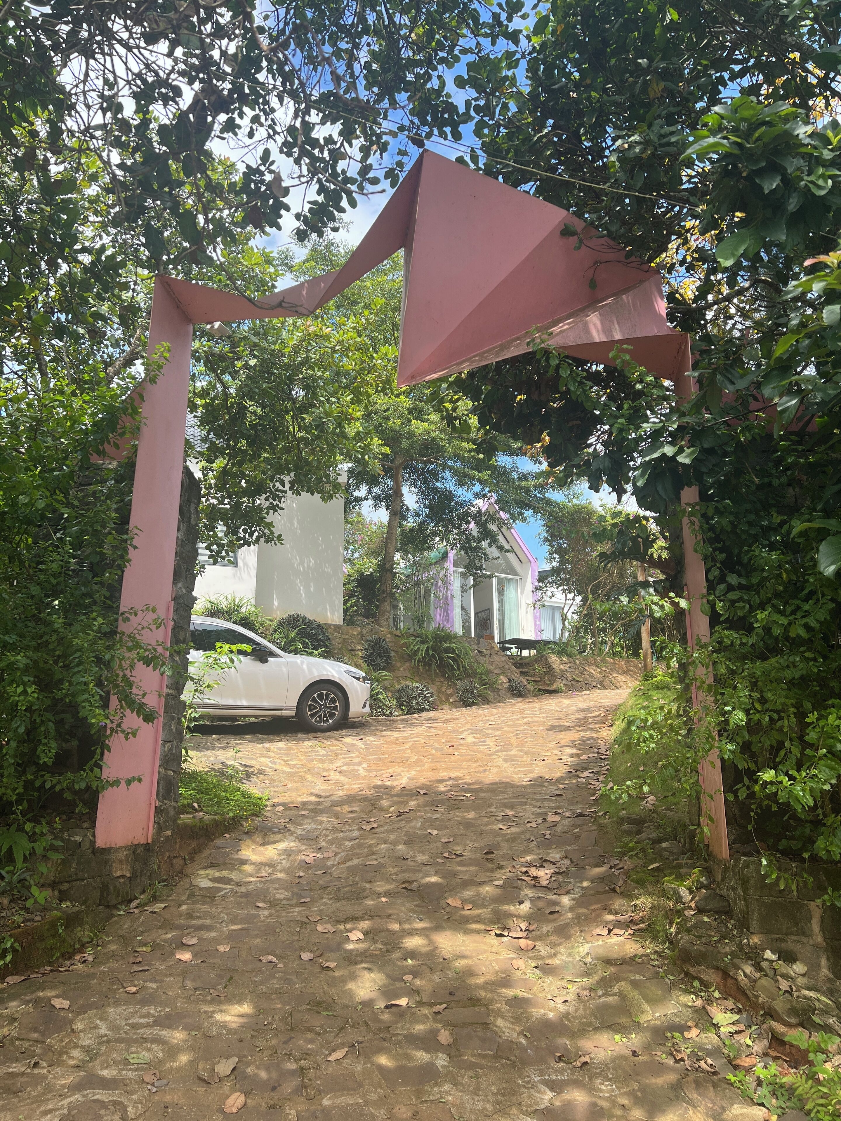 A paved pathway leading to a building entrance, surrounded by greenery and a white car parked nearby.